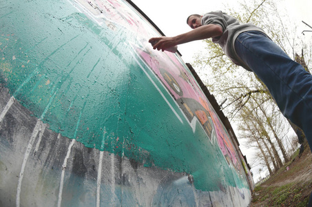 一个穿灰色连帽衫的年轻人在雨天在墙上画粉红色和绿色的涂鸦。 菲什耶枪