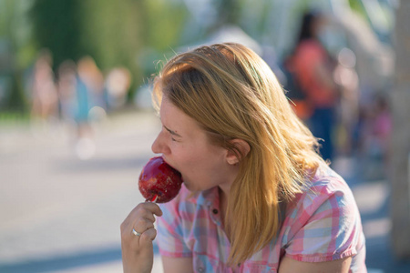 在阳光明媚的日子里，漂亮的年轻女人在公园里吃和咬红焦糖苹果
