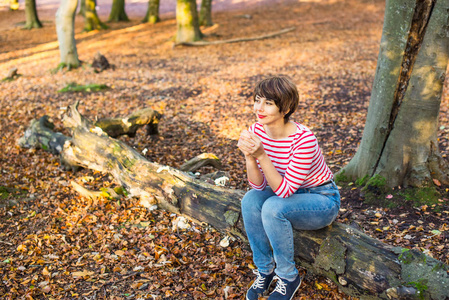 秋林中的年轻女子坐在倒下的树上放松的肖像。 把目光移开。 微笑的女孩。 秋天的户外生活方式。 与自然的平静和统一。 选择性聚焦复