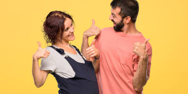 一对怀孕的夫妇竖起大拇指，微笑着，因为在孤立的黄色背景上取得了成功