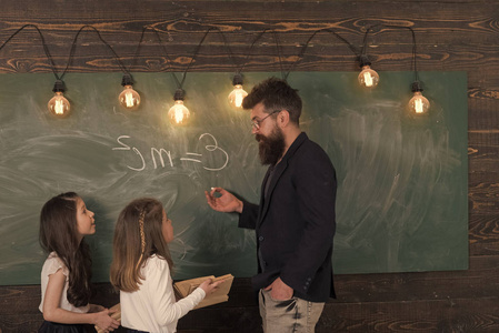 老师和女生学生在教室附近黑板。穿正式西装留胡子的男人教女学生物理。好奇的集中的孩子听老师的注意。物理课概念