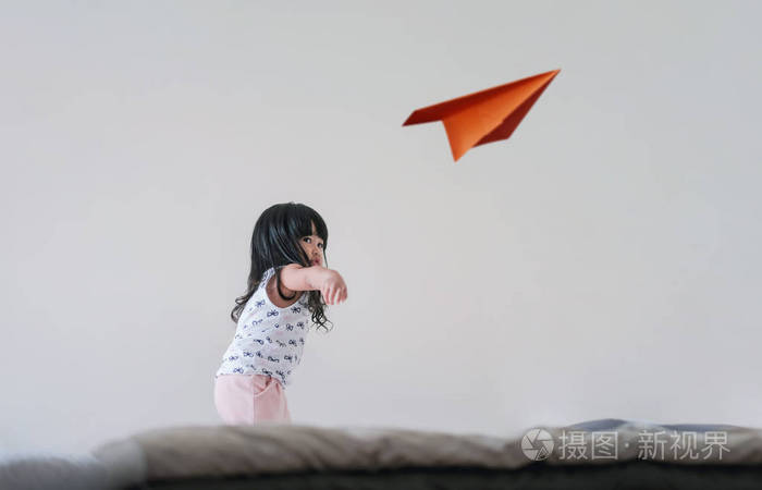 3岁的女孩举起手,在白色的房间里扔了一架纸飞机