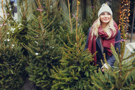漂亮的金发女孩在户外的树店里寻找完美的圣诞树。 女孩用手检查质量复制空间图像