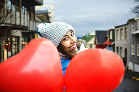 穿着温暖的冬衣针织帽子和围巾的快乐年轻女子站在户外, 带着心形的红色气球
