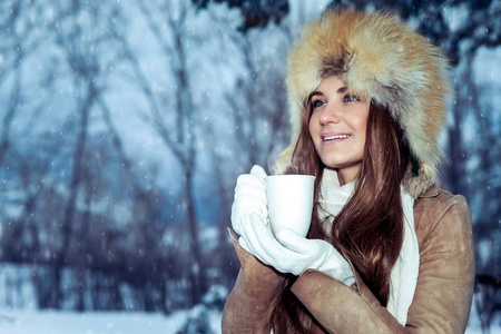 一位美丽的年轻女孩的肖像，戴着一顶时髦的帽子，站在一个下雪的公园里，温暖的双手放在一个热茶杯上，享受着美丽的景色