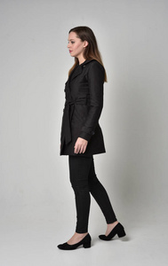 一个黑发女孩穿着长长的黑色外套，站在灰色工作室背景的侧面轮廓中的长度肖像。