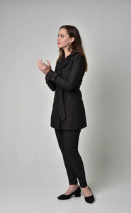 一个黑发女孩穿着黑色长外套，站在侧面轮廓灰色工作室背景的完整长度肖像。