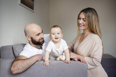 微笑的母亲和父亲抱着他们的女婴女儿坐在家里的床上。快乐的家庭概念, 穿着白衬衫的胡子男人, 快乐的女人抱着孩子