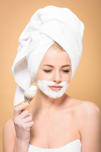 女人头上戴着毛巾，脸上戴着剃须膏，手里拿着剃须刷，低头看着米色的东西