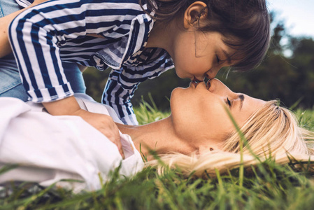 侧视特写肖像美丽的女人玩她可爱的小女孩在绿草户外。 爱快乐美丽的母亲亲吻她的女儿，花时间在公园里。 快乐的母亲