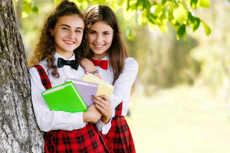 两个穿着红色学校格子制服的女学生站在靠近一棵树的公园里，那里有书