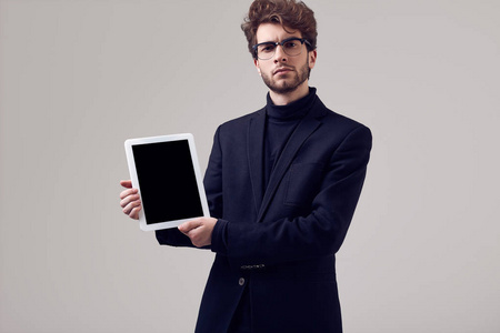 时尚肖像，英俊优雅的男人，卷发，穿西装，戴眼镜，在工作室里戴着一块灰色背景的平板电脑