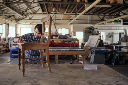 熟练的家具制造商在他的大型木工车间独自工作时，在工作台上打磨一把木椅