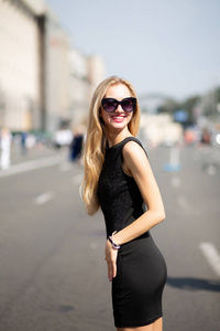 美丽的微笑的女人穿着黑色的裙子和太阳镜走在阳光明媚的街道上