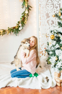 一位金发可爱的女孩在家里和一只小马拉莫特小狗玩耍，房间里装饰着圣诞节