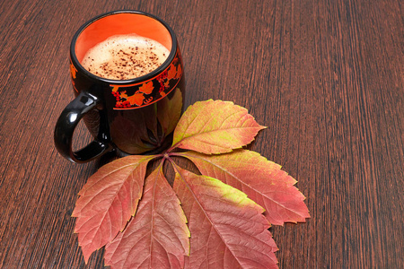 一杯泡沫咖啡放在一张棕色的木桌上，上面放着一片黄色的秋叶