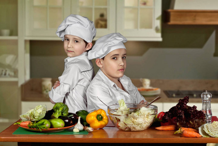 穿着厨师服装的男孩在厨房切沙拉。孩子们准备健康的食物。厨师的友好合作。
