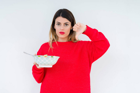 年轻女性在孤立的背景下吃新鲜的健康沙拉，愤怒的脸，负面的迹象，表示不喜欢，拇指向下拒绝的概念