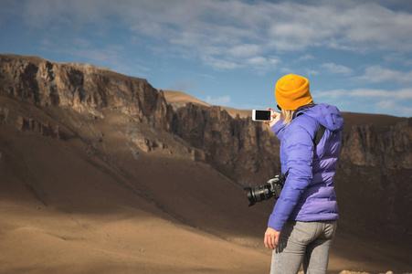 一位身穿羽绒服的旅行女孩的背部肖像, 她的帽子拍摄了一个史诗般的风景, 智能手机上有岩石
