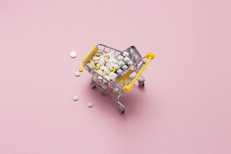 购物手推车从超市装满药丸和药物的粉红色背景。 在互联网上购买医疗制剂。 平躺式顶部视图
