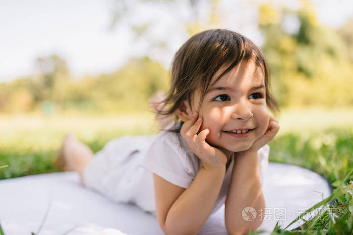 一个微笑可爱的小女孩躺在公园草地上的白色毯子上的水平图像.