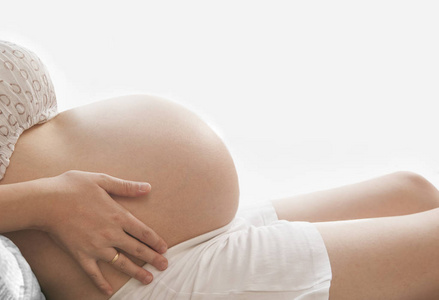 无法辨认的孕妇靠近白色窗户背景的腹部。 怀孕在家里的沙发上。 没有脸。