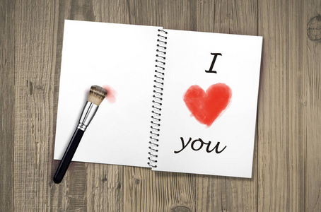 在老式木桌上打开的笔记本上，我爱你手写体和红色的心形，上面画着化妆画笔，作为庆祝浪漫情人节的信息