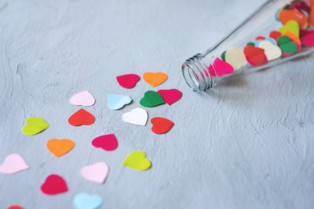情人节创意DIY工艺礼品卡的想法。 许多彩色纸心是从玻璃透明瓶在灰色水泥背景上倒入的。 最小爱情浪漫手工制作