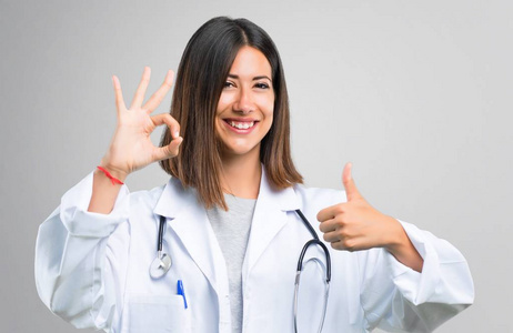 医生女士用听诊器显示一个确定的标志用手指和一个拇指向上手势与另一只手在灰色背景