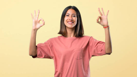 年轻女孩穿着粉红色的衬衫，手指上有一个OK的标志。 黄色背景下的幸福和满足感