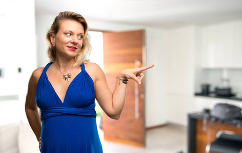 一位身穿蓝色连衣裙的金发孕妇，手指指向侧面，在她的房子里展示一种产品