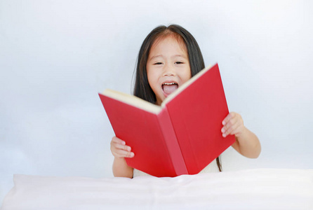 微笑的亚洲小女孩读精装书，躺在床上枕头在白色背景。