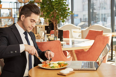 饥饿的头脑。 一位英俊的适合做生意的商人在餐馆用笔记本电脑吃饭