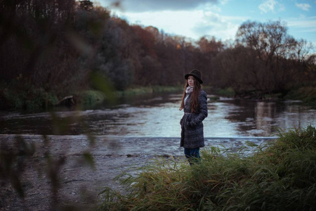 一个女孩在阴天的晚上站在桥附近的河岸上。 穿外套戴帽子的年轻女子站在水边
