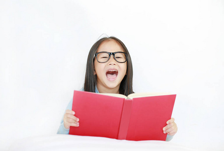 微笑的亚洲小女孩戴着眼镜，在白色背景下躺在床上看书。