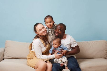 幸福的多民族家庭的肖像坐在家里的沙发上看着相机。 黑人父亲，白人母亲和两个女儿。 拥抱对方。