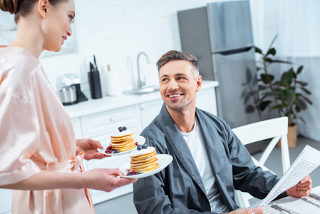 女人拿着煎饼盘子，微笑着男人在厨房吃早餐时看报纸