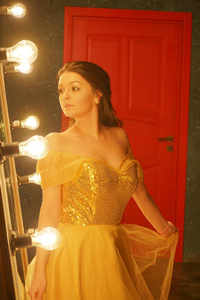 一个穿着金色晚礼服的漂亮少女站在一块毛地毯上，靠近一面大镜子，镜框里有灯光，看着她的倒影