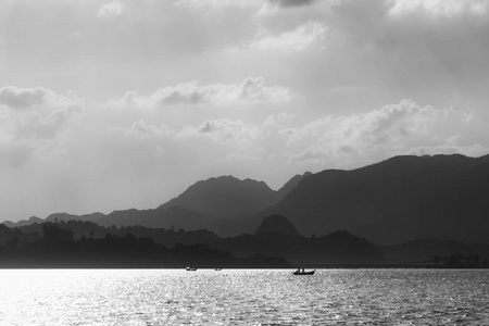 在群山的背景下，渔船的轮廓和一个晴朗的天空与云。 黑白摄影