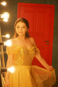 一个穿着金色晚礼服的漂亮少女站在一块毛地毯上，靠近一面大镜子，镜框里有灯光，看着她的倒影