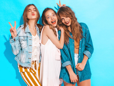 三个年轻美丽的微笑时髦的女孩穿着时髦的夏季服。 无忧无虑的女人在蓝墙附近摆姿势。 积极的模特发疯了
