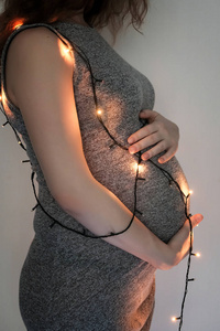 一个穿灰色衣服的年轻怀孕女孩抱着她的肚子，周围挂着灯。 新年和圣诞节的概念。