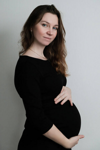 穿着黑色衣服的年轻怀孕女孩站在白色背景上拥抱她的腹部。