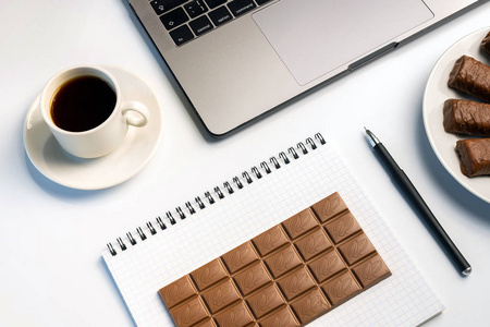 在工作中吃零食有一个咬人的概念。 笔记本电脑糖果和工作簿与巧克力和咖啡在工作场所。 平躺顶部视图白色桌子。