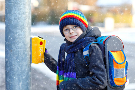 在降雪期间, 小学的小学生走着去上学。快乐健康的孩子用眼镜按下红绿灯的按钮。用背包或背包穿着五颜六色的冬衣