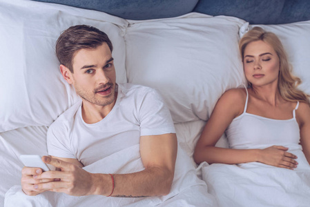 年轻人使用智能手机和看相机的高角度视角，与熟睡的妻子躺在床上