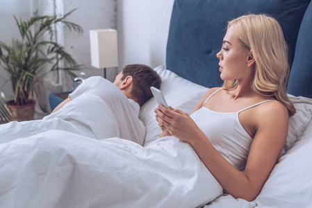嫉妒的年轻女人使用智能手机，看着男朋友睡在床上，不信任概念