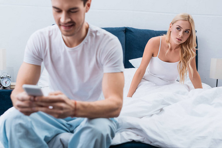嫉妒的年轻女人在卧室的前台看男朋友用智能手机