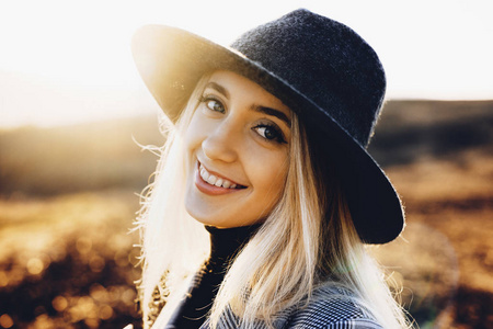 美丽的年轻女子戴着时尚的帽子，兴高采烈地微笑着，看着摄像机，站在阳光明媚的秋天自然的模糊背景上。农村的善良女性