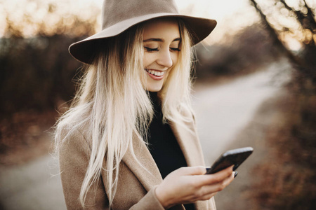 迷人的年轻女子戴着优雅的帽子，兴高采烈地微笑着，一边使用智能手机，一边站在模糊的秋天乡村背景上。潇洒的女士浏览大自然中的智能手机